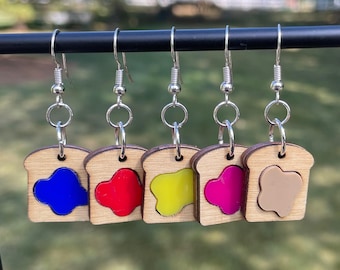 May include: Five pairs of earrings shaped like slices of bread with different colored jelly fillings. The fillings are blue, red, yellow, pink, and brown.