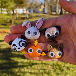 May include: Five colorful googly-eyed animal figurines, including a white bunny, a black and white panda, a brown and white pug, a brown and white cat, and an orange fish.