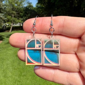 May include: A pair of silver-toned dangle earrings. Each earring features a clear, rectangular pendant with a blue and gold design, accented by red and pink lines. The earrings are suspended from silver-colored hooks.