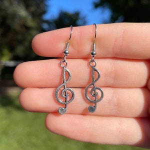 May include: A pair of silver-toned earrings featuring a treble clef design. The earrings have a simple hook closure and a small, round bead detail. The background is a blurred view of green foliage and blue sky.