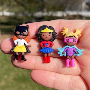 May include: Three small plastic superhero figurines. The first figurine is a black and yellow batgirl with a white shirt and a yellow skirt. The second figurine is a Wonder Woman with a red shirt, blue skirt, and a gold headband. The third figurine is a purple superhero with a blue cape and pink boots.
