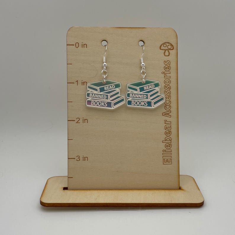 May include: A pair of iridescent blue and green dangle earrings featuring a stack of books with the words "Read", "Banned", and "Books" on them. The earrings are displayed on a wooden stand with a ruler in inches.