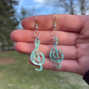 May include: A pair of silver dangle earrings with a light blue treble clef charm. The charms are filled with iridescent glitter.