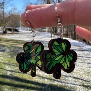 May include: A pair of wooden shamrock earrings with silver-colored hooks. The shamrocks have a dark brown border and a stained-glass effect with shades of green and yellow.