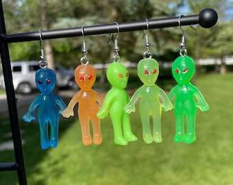 Peut inclure: Cinq boucles d'oreilles en forme d'extraterrestre, chacune d'une couleur translucide différente : bleu, orange, vert, vert-jaune et vert. Chaque extraterrestre a des yeux rouges et est suspendu à un crochet argenté. Les boucles d'oreilles sont exposées sur un fond vert flou.