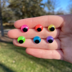 May include: Six colorful googly eyes with black pupils. The eyes are arranged in a semi-circle on a hand.