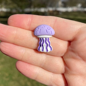 May include: A small, purple and white jellyfish charm. The charm has a rounded top with small circles and lines, a white band, and wavy purple and white tentacles. The charm is held in a hand.