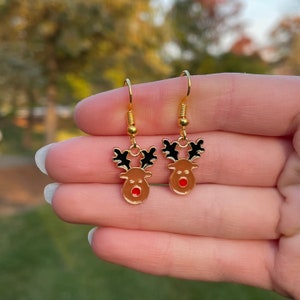 May include: A pair of gold-colored dangle earrings featuring reindeer designs. Each earring has a brown reindeer head with black antlers and a red nose. The earrings are held up by gold-colored hooks.