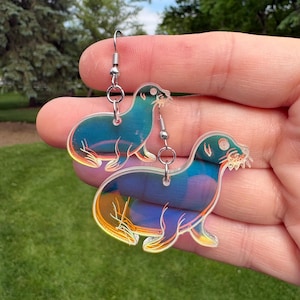 May include: A pair of iridescent seal-shaped earrings. The earrings feature a holographic effect with colors of blue, purple, and gold. Each earring has a silver-colored fish hook and a small silver bead.
