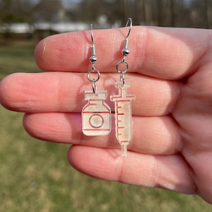 Peut inclure: Une paire de boucles d'oreilles en argent avec des breloques en acrylique transparent. Un charme est un flacon avec un bouchon rouge et l'autre est une seringue.