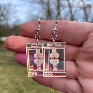 May include: A pair of iridescent earrings with a design of a computer screen displaying a pair of bones. The earrings are silver and have a hook closure.