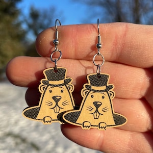 May include: A pair of wooden earrings featuring a cartoon groundhog wearing a top hat. The groundhog is depicted emerging from a hole in the ground. The earrings are made of natural wood and have a black and brown colour scheme.