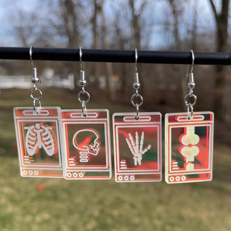 May include: Four pairs of earrings with a red and white holographic effect. Each earring features a different medical x-ray image: a chest x-ray, a skull x-ray, a hand x-ray, and a spine x-ray.