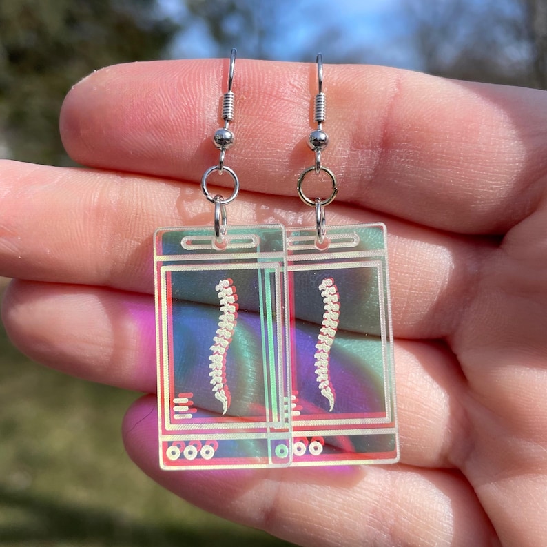 May include: A pair of silver earrings with clear acrylic charms featuring a spine x-ray image. The charms have a holographic iridescent finish.