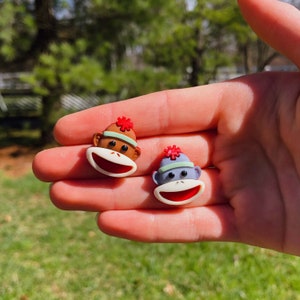 May include: Two small monkey-shaped buttons with smiling faces. One is brown with a red hat and the other is gray with a blue hat.