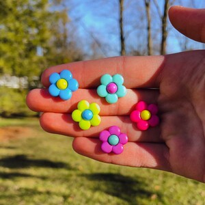 May include: Five colorful flower-shaped buttons in shades of blue, green, pink, and yellow. The buttons are arranged on a hand.