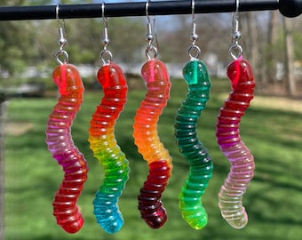 May include: Five colorful gummy worm earrings hanging from a black rod. Each earring features a different color gradient, including red, orange, yellow, green, and purple. The earrings have silver-colored hooks.
