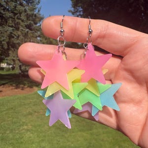 May include: Dangle earrings featuring multiple layered star-shaped charms in translucent neon colors including pink, yellow, green, and purple. The stars are suspended from silver-toned metal hooks and jump rings, creating a cascading effect.