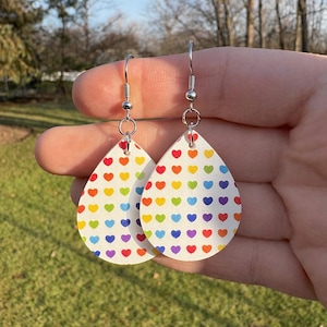 May include: Teardrop-shaped earrings with a white background and a pattern of colorful hearts. The earrings have silver-colored hooks. The hearts are in a rainbow of colors, including red, orange, yellow, green, blue, and purple.