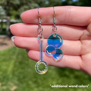 May include: Iridescent bubble-themed earrings. One earring features a bubble wand design, while the other has a cluster of three bubbles. The earrings are suspended from silver-colored ear wires. The text "additional wand colors" is at the bottom.
