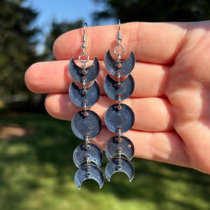 May include: Dangling earrings featuring a celestial design. Each earring showcases a series of dark blue, crescent moon and circular shapes connected by silver-toned metal. The earrings have a speckled, glittery appearance, adding a touch of sparkle.
