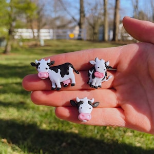 May include: Three black and white cow figurines with pink noses. The cows are standing on a hand.