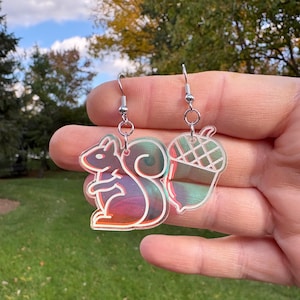 May include: A pair of dangle earrings featuring a squirrel and an acorn. The squirrel has a red, purple, and green iridescent finish with a white outline. The acorn has a green and brown iridescent finish with a white grid pattern.