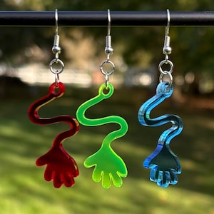 May include: Three pairs of dangle earrings in red, green, and blue. Each earring has a wavy, elongated design with a hand-shaped end. The earrings are suspended from silver-coloured hooks, creating a playful and colourful accessory.
