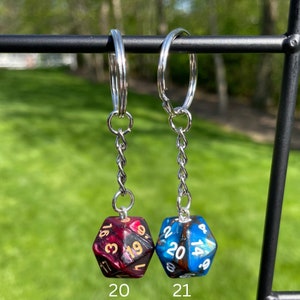 May include: Two silver keychains with a black and red dice charm on the left and a blue and black dice charm on the right. The dice charms are numbered 1-20. The keychains are hanging from a black metal bar.