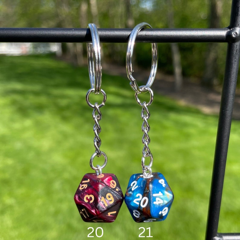 May include: Two silver keychains with a black and red dice charm on the left and a blue and black dice charm on the right. The dice charms are numbered 1-20. The keychains are hanging from a black metal bar.