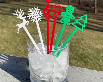 May include: Six festive acrylic drink stirrers in a glass filled with ice. The stirrers feature a reindeer, snowflake, the word "JOY", a candy cane, a gingerbread man, and a Christmas tree. The stirrers are white, red, and green.