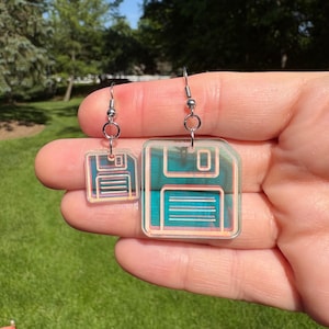 May include: A pair of clear acrylic earrings shaped like retro floppy disks. The earrings have a teal and pink color scheme with silver-colored ear wires. The design features the iconic square shape and details of a classic 3.5-inch floppy disk.