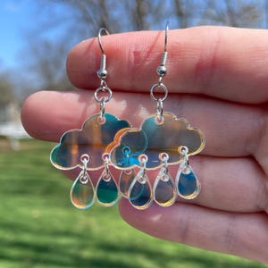 May include: A pair of iridescent silver earrings featuring a cloud with raindrops hanging from it. The earrings are made of clear acrylic and have a holographic finish.