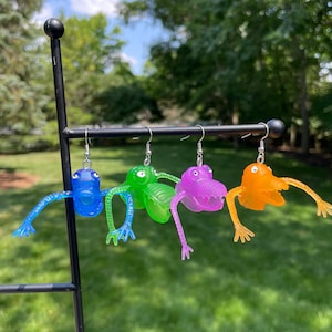 May include: Four pairs of novelty earrings in blue, green, purple, and orange. Each earring features a cartoon-like creature with outstretched arms and legs, suspended from a silver hook. The earrings are displayed on a black metal stand against a blurred green background.