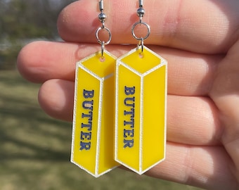 May include: A pair of yellow stick-shaped earrings with the word "BUTTER" printed on each. The earrings are made of acrylic and have silver hooks.