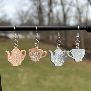 May include: Four pairs of dangle earrings featuring tea-themed designs. The earrings include two teapots and two teacups, each with a floral pattern. The colors are peach, tan, and gray, with white detailing. The earrings hang from silver-colored hooks.