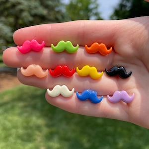 May include: Assortment of ten colorful plastic mustache shaped buttons. The buttons are in the following colors: pink, green, orange, peach, red, yellow, black, white, blue, and purple.