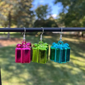 May include: Three earrings shaped like gift boxes in magenta, lime green, and teal. Each has a bow and a silver hook. The earrings are suspended against a blurred green backdrop.