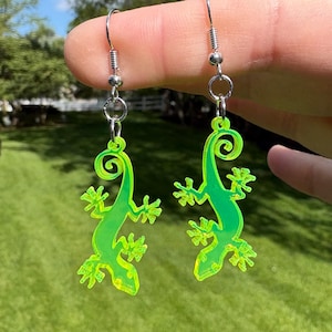 May include: A pair of neon green gecko earrings. Each earring features a stylized gecko shape with a curled tail and detailed feet. The earrings are suspended from silver-colored fish hook ear wires.