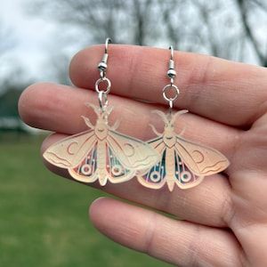 May include: A pair of moth-shaped earrings with silver-colored hooks. The earrings feature a cream-colored body with red, blue, and green wing details. The moth design includes crescent moon shapes and circular patterns.