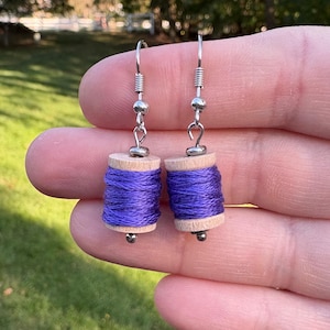 May include: A pair of handmade earrings featuring miniature wooden spools wrapped with vibrant purple thread. The earrings have silver-colored hooks and small silver beads. The spools are approximately 0.75 inches in height.