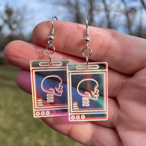 May include: A pair of iridescent earrings featuring a skull graphic inside a rectangular frame. The earrings are made of clear acrylic and have silver hooks.