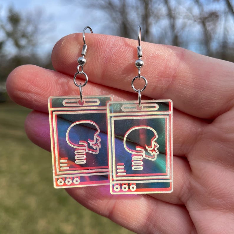 May include: A pair of iridescent earrings featuring a skull graphic inside a rectangular frame. The earrings are made of clear acrylic and have silver hooks.