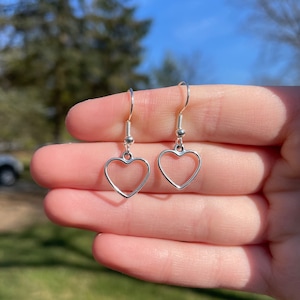 May include: A pair of silver heart-shaped dangle earrings. Each earring features a heart outline suspended from a silver hook. The earrings are held up against a blurred green and blue background.
