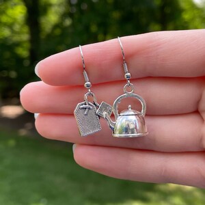 May include: Silver-toned dangle earrings featuring a tea kettle charm and a tea bag tag charm. The earrings have a simple hook design and are likely made of a lightweight metal. The background is blurred, suggesting an outdoor setting.