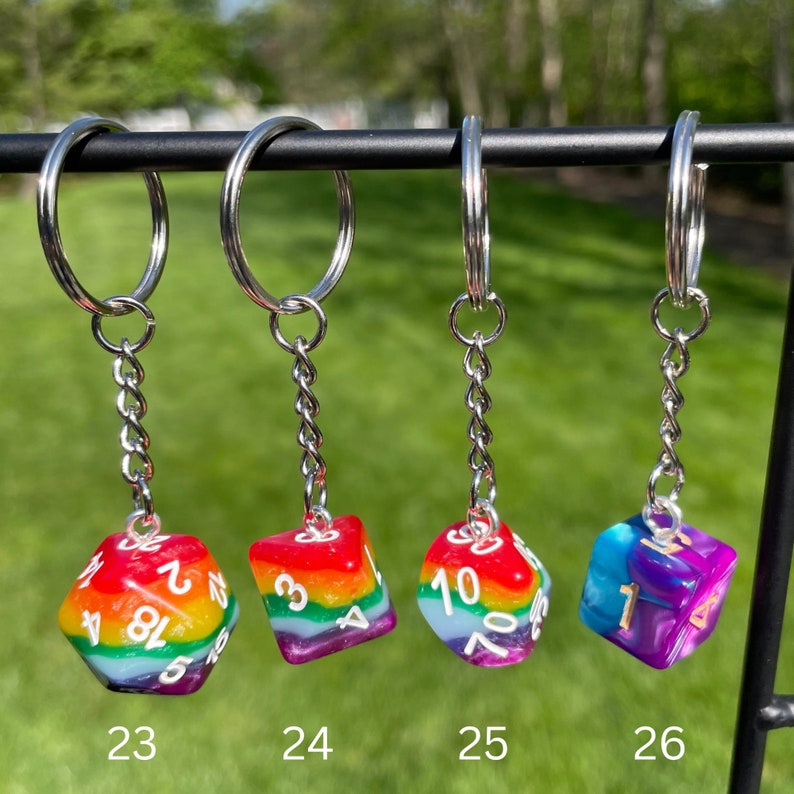 May include: Four rainbow dice keychains with silver key rings and chains. The dice are numbered 1-6 and have a rainbow swirl pattern. The dice are labeled 23, 24, 25, and 26.