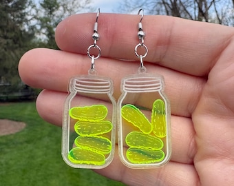 May include: A pair of novelty earrings featuring clear acrylic jars filled with neon green pickle shapes. The earrings have silver-colored fish hook ear wires. The background is a blurred outdoor scene with green grass and trees.