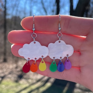 May include: A pair of silver dangle earrings featuring a white cloud charm with rainbow raindrop charms hanging below.