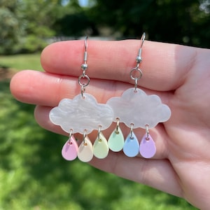 May include: A pair of cloud-shaped earrings with pastel-colored raindrop charms. The earrings feature a silver hook and are held in a hand against a blurred green background. The raindrops are pink, yellow, green, blue, and purple.