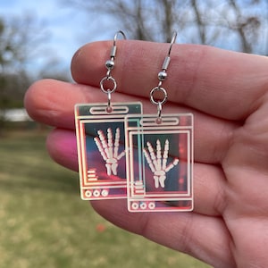 May include: A pair of iridescent silver earrings with a holographic finish. Each earring features a rectangular pendant with a white outline of a hand x-ray image.